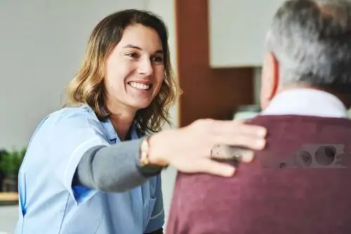 Zorgprofessional in lichtblauw shirt geeft een oudere patiënt een geruststellende handdruk in een huiselijke omgeving.